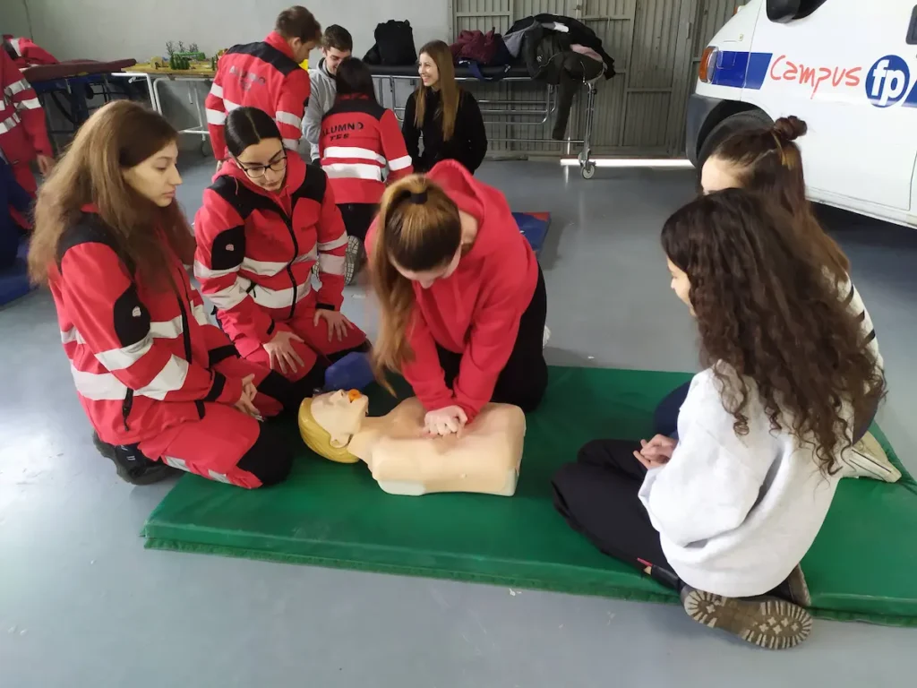 Jóvenes sobre una colchoneta verde practicando reanimación cardiopulmonar en un maniquí de entrenamiento médico.